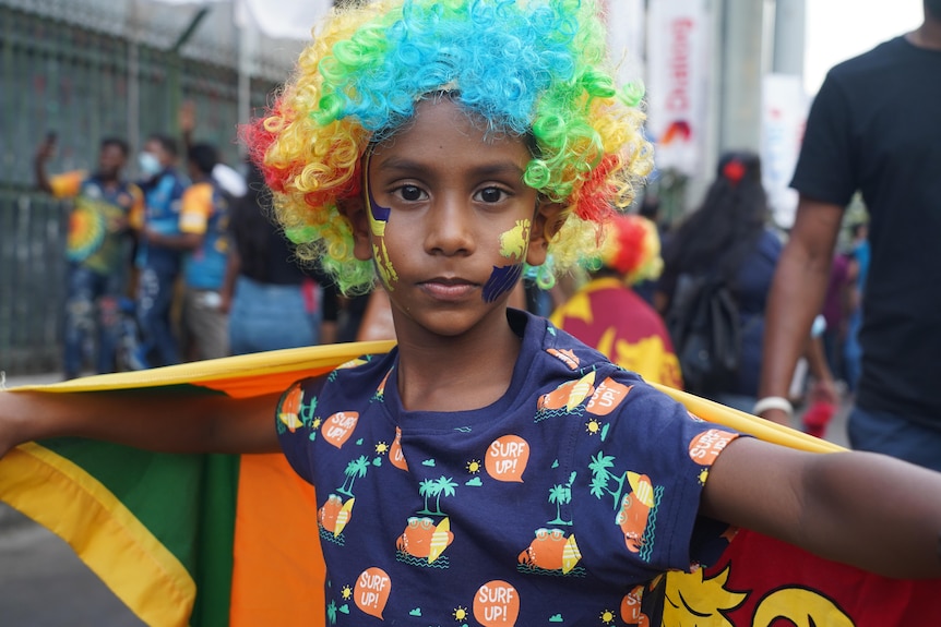 A young boy in a curly clown wig holds a Sri Lankan flag around him 