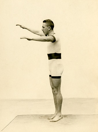 Old sepia photo with side profile of man in full-body swimming outfit, who has arms out-stretched as if about to dive into pool.