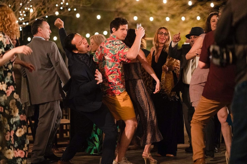 Andy Samberg holding up a drink and dancing in a wedding crowd in a scene in the film Palm Springs