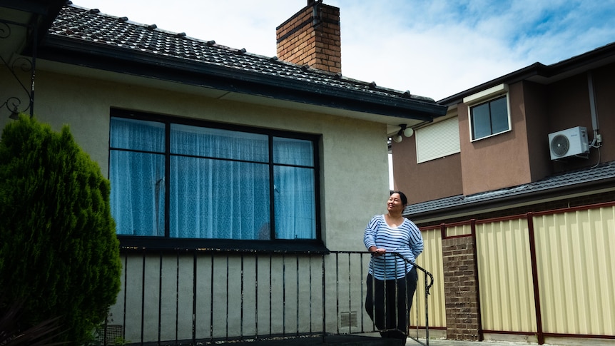 A woman with dark hair and a striped shirt gazing from her front verandah off camera.