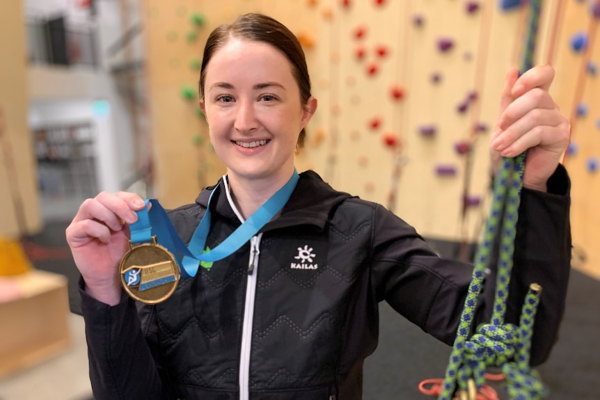 Sarah Larcombe smiles as she holds up a gold medal around her neck in one hand and climbing ropes in the other.