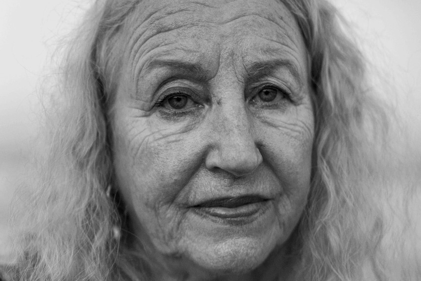 An older woman with curly white hair stares into the camera looking sombre.