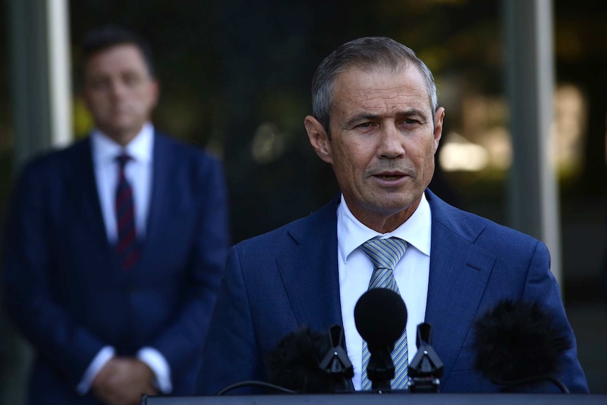 WA Health Minister Roger Cook, wearing a blue suit, speaking into a microphone at a press conference