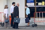 Four people outside a sliding door, one older man in a suit carrying a briefcase, a younger man next to him, and two women