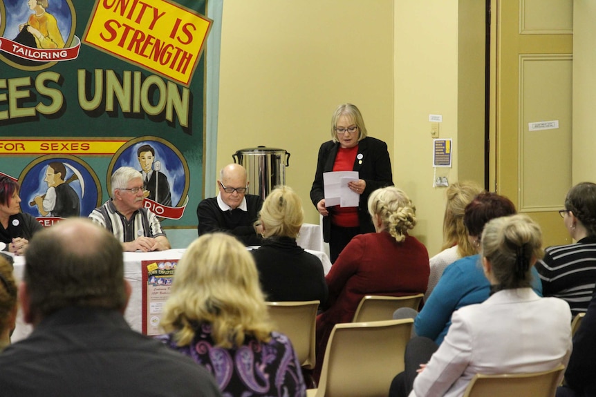 Wide-shot of Maureen stands and looks at sheet of paper while she addresses public.