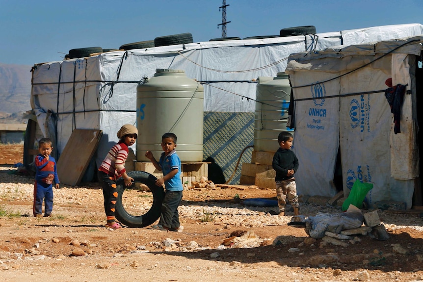 Children play with a tyre in a refugee camp with water tanks and makeshift tents with the UNHCR logo.