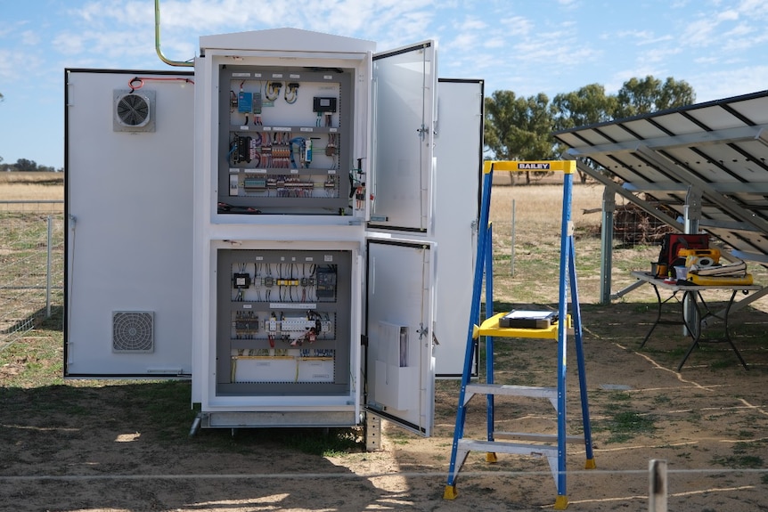 A large white box with exposed wiring beside a solar panel