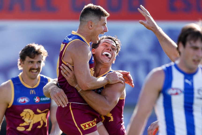 Two Brisbane Lions players yell and grin in celebration after a goal, as a Kangaroos defender walks away.