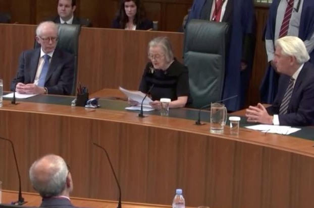 Two men sat either side of Lady Brenda Hale look to her as she reads from a piece of paper sat at a brown desk.