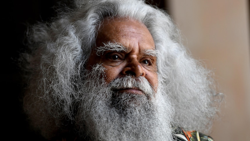Uncle Jack Charles looks thoughtful, standing in gentle light.