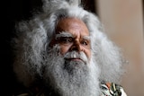 Uncle Jack Charles looks thoughtful, standing in gentle light.