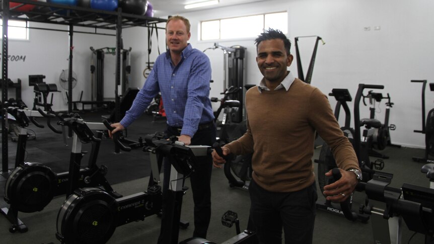 President Carl Semler is standing inbetween spin bike wearing a blue sit. Next to him is committee member Rav Prakash. 