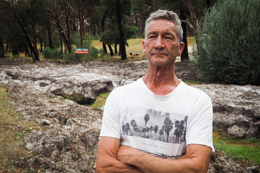 A man in a white-t-shirt stands in front of a roadside coated in hail.