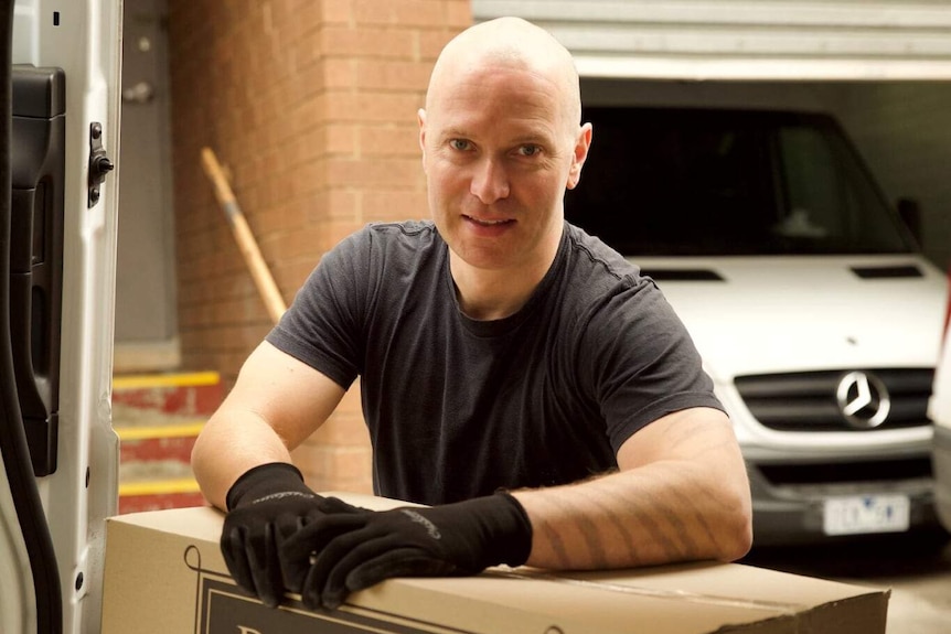 A man in a black shirt and gloves looks at the camera while resting on a box in a van.