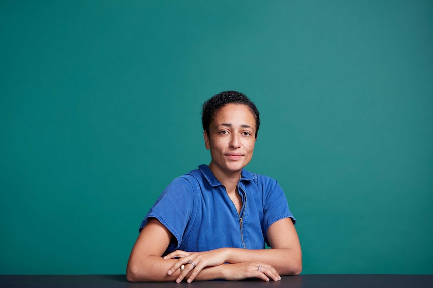 The writer Zadie Smith with a serious look on her face, arms in front of her, teal background