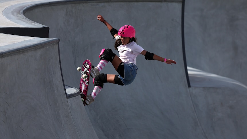 Arisa Trew on a skating ramp wearing a pink helmet with her arms spread out standing on an angle on a skateboard.