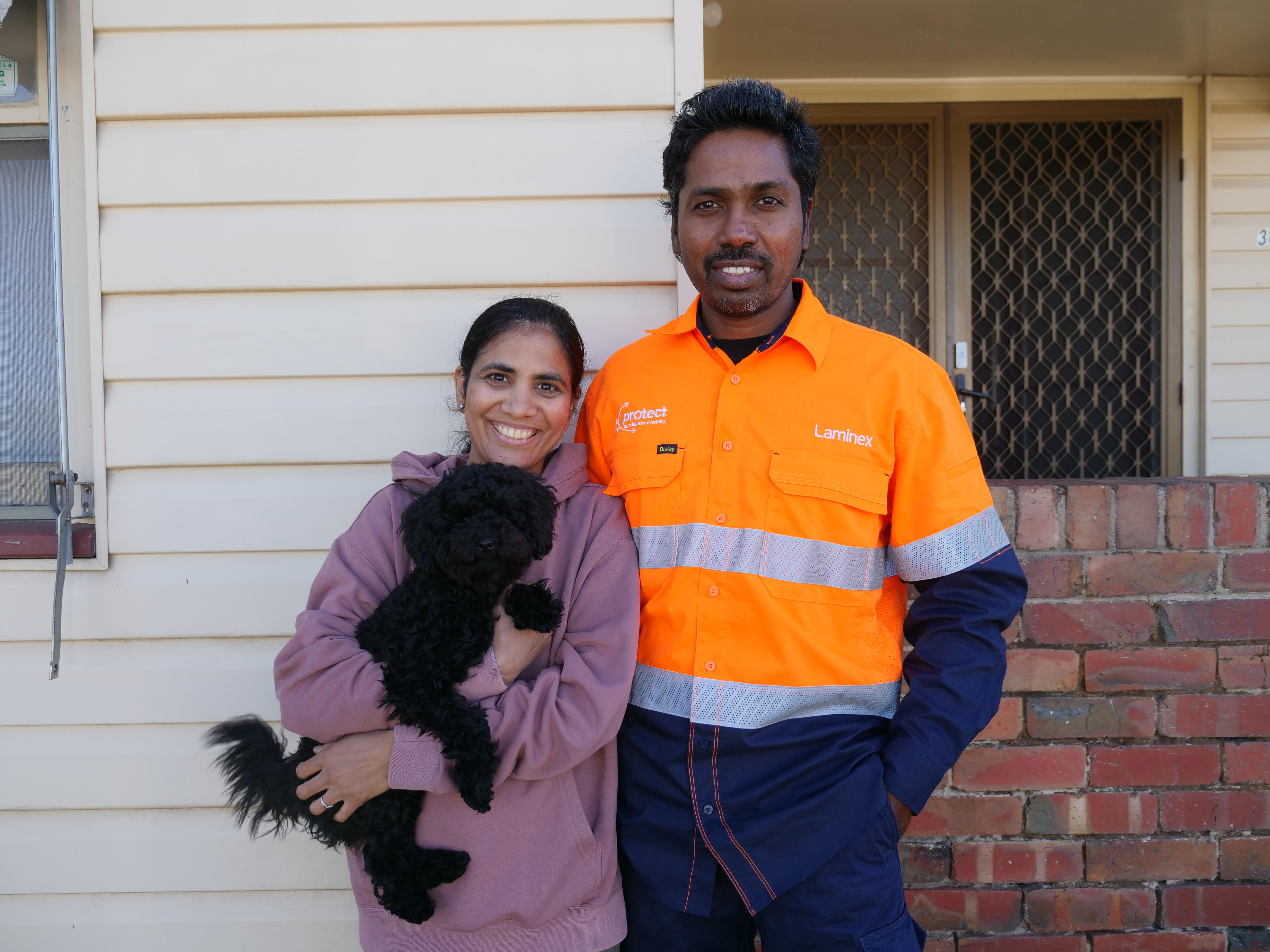 A brown skinned woman and man stand next to each other outside a weatherboard house, the woman is holding a black dog. 
