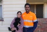 A brown skinned woman and man stand next to each other outside a weatherboard house, the woman is holding a black dog. 