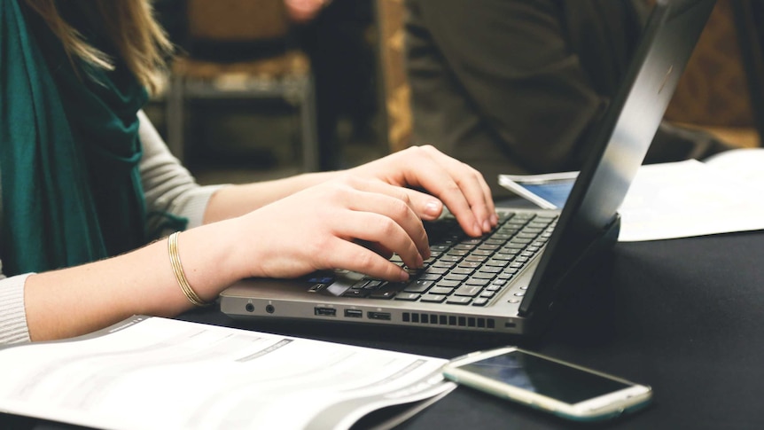 Two hands typing on a laptop with a phone and paper in the foreground