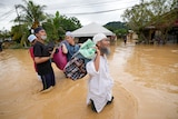 Five men wearing islamic headdress wade through knee high brown floodwaters lugging bags of medical supplies.