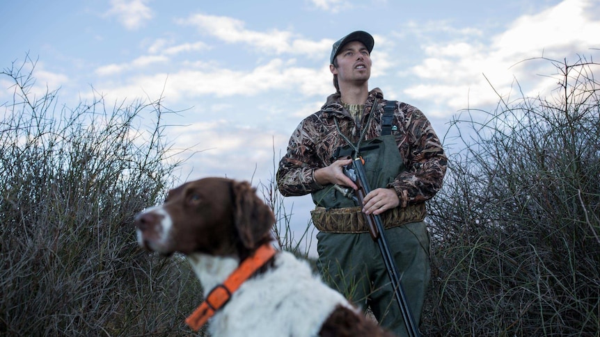 Dressed in camouflage gear and clutching his gun, duck hunter Dean Rundell looks skyward, flanked in the marsh by his dog.