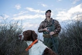 Dressed in camouflage gear and clutching his gun, duck hunter Dean Rundell looks skyward, flanked in the marsh by his dog.
