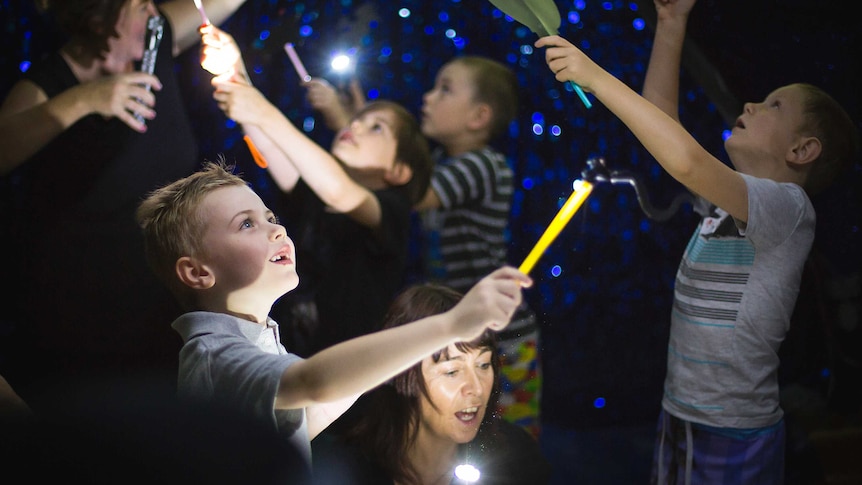 A fascinated child points a yellow stick up to the ceiling, illuminated by a performer's torch.