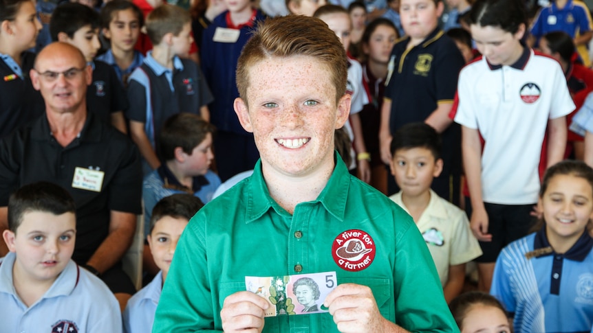 A boy stands in front of a large crowd, holding a $5 note.
