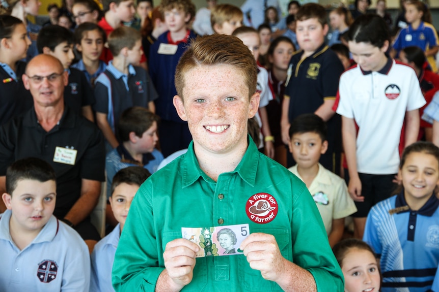 A boy stands in front of a large crowd, holding a $5 note.