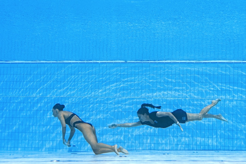 A woman swims out to grab another unconscious woman at the bottom of a pool.