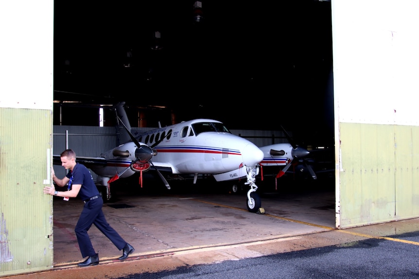 A man pushes open a rolling door, revealing a small plane inside the hangar. 