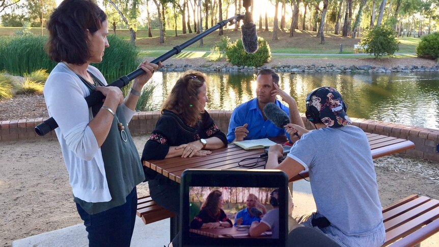 Producer holding microphones as Clarke and Smith talk while sitting at picnic table next to river