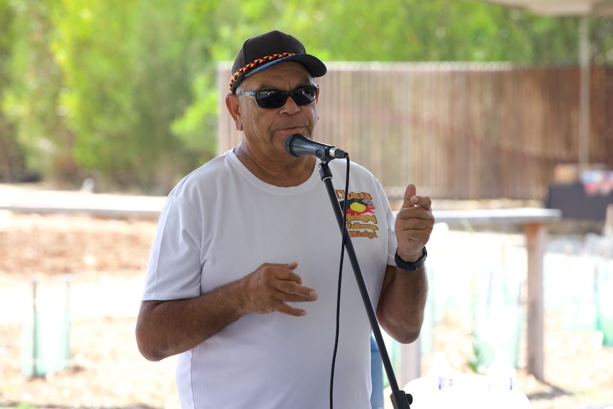An Indigenous man wearing sun glasses and a hat speaks at a mircophone.