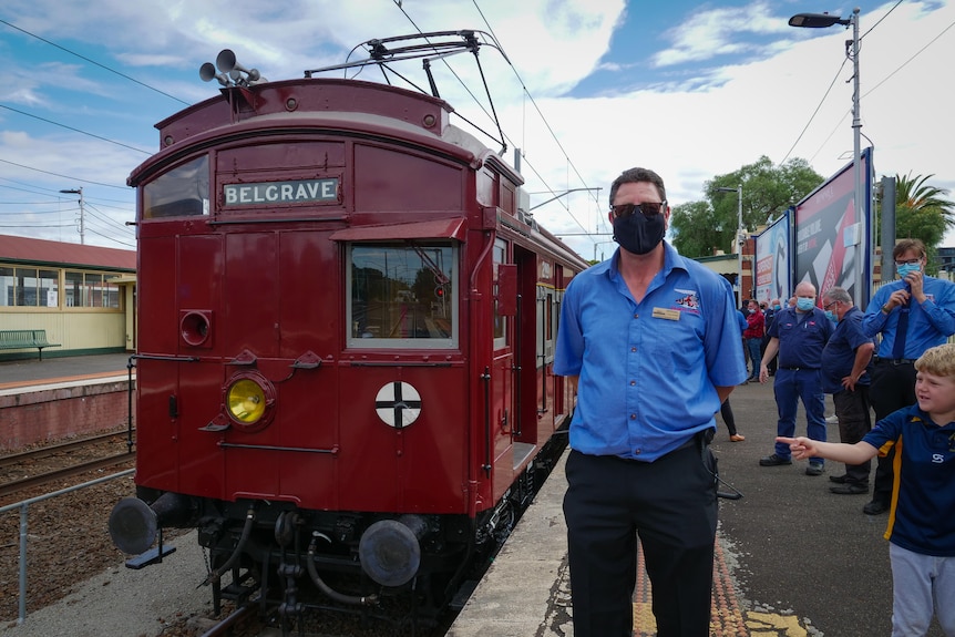 A man in uniform stands in front of a beautifully restored heritage red train with 'Belgrave' written on the front.