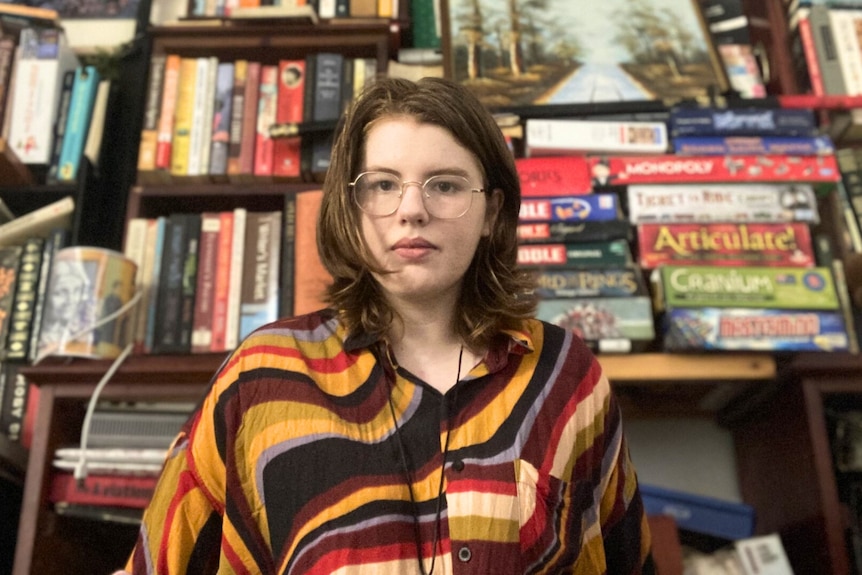 A young white woman with glasses and brown hair wearing a colourful shirt standing in front of a bookcase