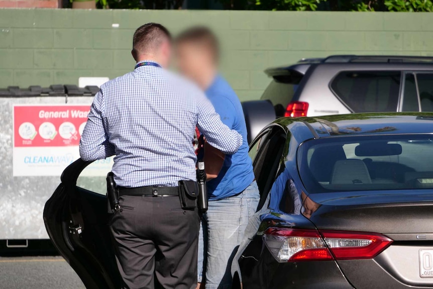 A man with his face blurred leaving an unmarked police car next to a plain-clothed detective