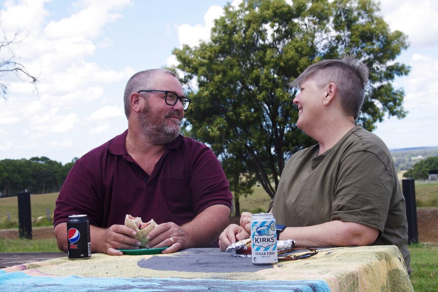 A man and a woman sitting at a picnic table holding food smile at each other