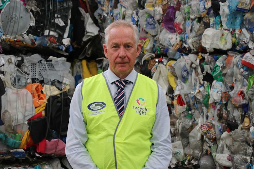A man stands in a shirt, tie and fluorescent vest against a wall of compacted recycling.