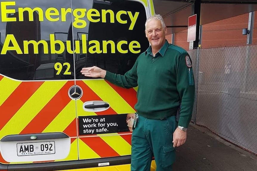 Paramedic in green work clothes with grey hair stands next to ambulance