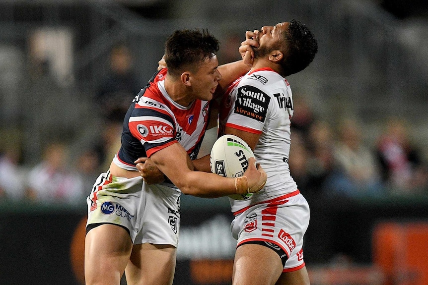 A male NRL player holds the ball with his right hand as he palms off an opponent in the face with his left hand.