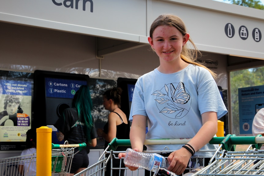 A teenage girl ina  bright blue shirt holding an empty plastic water bottle in front of a Return and Earn depot.