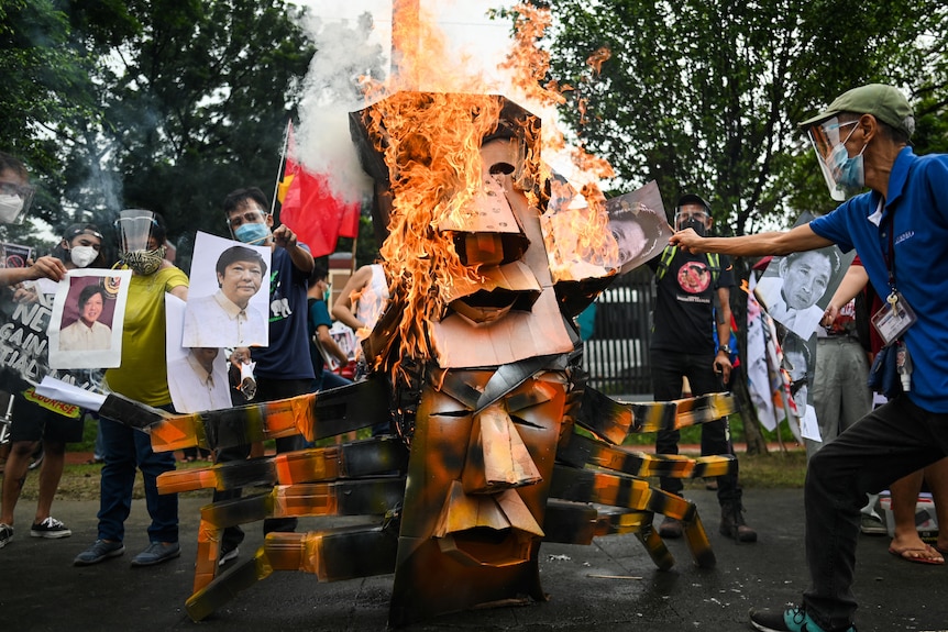 A two-headed effigy depicting Rodrigo Duterte and Ferdinand Marcos Jr is burned in the streets by protesters holding photos.