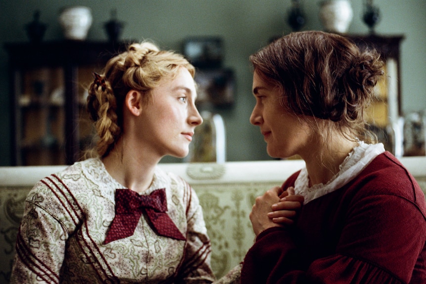 Close-up shot of Winslet and Ronan in 19th century dresses sitting on couch close, looking lovingly at each other.