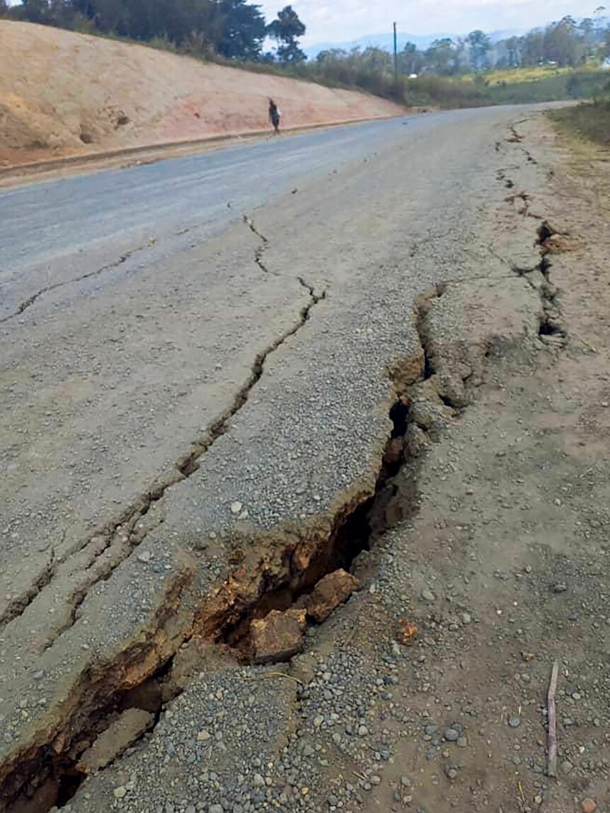 A large crack appears on a road. A man is walking in the background. 