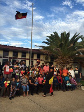 A group of protestors stand below a flagpole flying the Aboriginal flag outside the Shire of Carnarvon's offices.