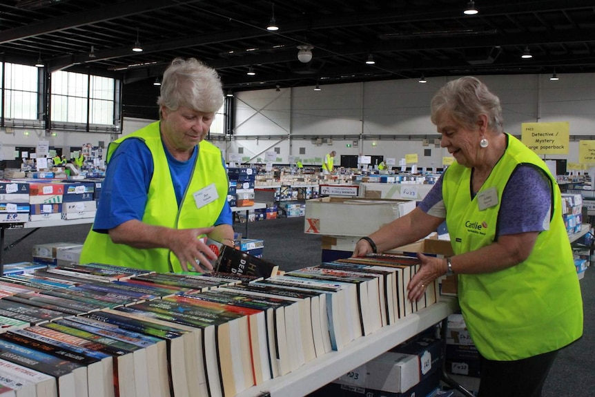 Two people wearing blue shirts and yellow vests look at row of books
