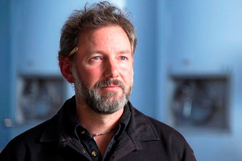 A close profile of a bearded, middle-aged Jeremy Dinnen. Background is blue and blurred.