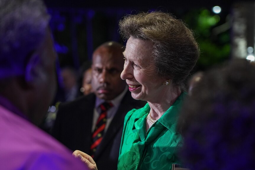 Princess Anne, in a green jacket, smiles as she chats with people at a night-time function