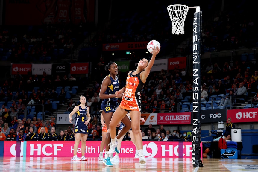 A Giants Super Netballer reaches out near the basket and catches the ball in the palm of her left hand as a defender trails. 