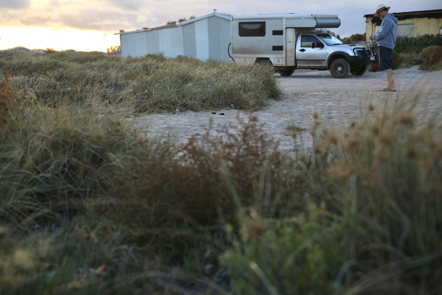 A sandy camp site at dusk, surrounded by beach scrub with a man and a vehicle and tin shacks in the background.